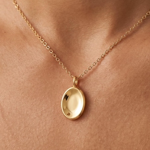 Puede incluir: Un collar con colgante ovalado dorado. El colgante tiene una forma ovalada lisa y pulida con un ligero diseño cóncavo. La cadena es una delicada cadena de eslabones dorados. El collar se muestra sobre un fondo color piel.