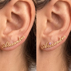 May include: A gold-toned ear climber earring featuring the name Alexandra in a cursive script font. The second version of the earring includes a small, round, pink gemstone accent positioned near the first letter of the name.