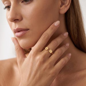 Könnte beinhalten: Ein goldfarbener offener Ring mit zwei kleinen runden Scheiben an den Enden. Jede Scheibe ist mit einem einzelnen Großbuchstaben graviert, den Buchstaben J und F, die so am Finger positioniert sind, dass die Initialen deutlich sichtbar sind.