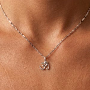 Puede incluir: Un collar plateado con un pequeño colgante con el símbolo Om. El colgante cuelga de una fina cadena. El collar se lleva alrededor del cuello.