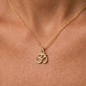 May include: A gold necklace with an Om symbol pendant. The pendant has a small, clear gemstone. The necklace is worn around the neck and is a delicate piece of jewellery.