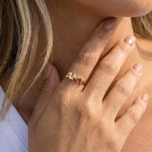May include: A gold ring spelling "AMY" in capital letters. The ring is worn on a finger, with the hand near a person's neck. The ring is the focal point, showcasing a personalized jewellery item. The ring is likely made of gold.