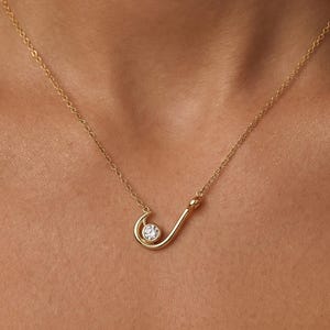 Könnte beinhalten: Eine goldfarbene Halskette mit einem geschwungenen Anhänger, der einen kleinen, runden, klaren Edelstein hält. Der Anhänger ist an einer filigranen Kette befestigt. Die Halskette wird um den Hals getragen.