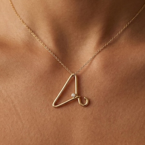 Puede incluir: Un collar dorado con un colgante con la letra "A". El colgante tiene un pequeño diamante. El collar se lleva alrededor del cuello.