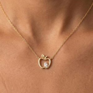 May include: A gold necklace with an apple-shaped pendant. The pendant has a leaf detail and a clear, oval-shaped stone. The necklace is displayed on a person's skin. The chain is delicate.