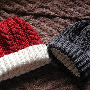 Può includere: Due berretti invernali lavorati a maglia. Uno è rosso con bordo bianco, l'altro è grigio scuro con bordo color crema. Entrambi i cappelli hanno un motivo a trecce. I cappelli sono esposti su una superficie marrone strutturata.