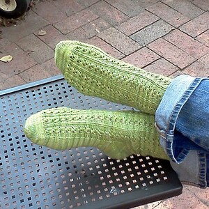 Könnte beinhalten: Ein Paar grüne Stricksocken mit einem Spitzenmuster. Die Socken werden an den Füßen einer Person getragen und liegen auf einem schwarzen Metalltisch.
