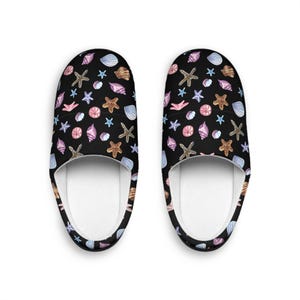 May include: A pair of black slippers featuring a repeating watercolor pattern of various seashells and starfish in shades of pink, blue, brown, and white. The slippers have a rounded toe design and a plain white interior lining visible from above.