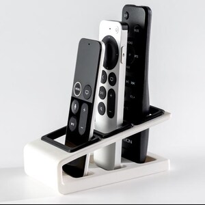 May include: A white and black remote control organizer holding three remotes. The remotes are black and silver. The organizer is white with a black accent.