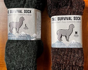 May include: Two pairs of The Survival Sock, crew socks made in the USA with U.S. grown alpaca. One pair is dark green, size L (10-13), and the other is brown, size M (7-10). Each sock has a label with an alpaca illustration.
