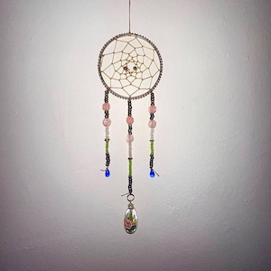 May include: A handcrafted dreamcatcher with a circular frame and a web design. The dreamcatcher features pink, green, and clear beads, with a small, oval pendant at the bottom. The pendant has a floral design and blue teardrop beads.