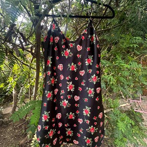 Op de afbeelding: Zwarte satijnen slipdress met een bloemen- en hartpatroon. De jurk heeft rode rozen en hartvormige ontwerpen met rode accenten. De jurk hangt aan een zwarte hanger tegen een achtergrond van groen gebladerte.