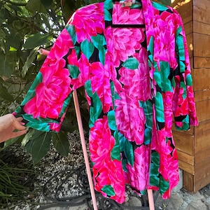 May include: A vibrant, open-front robe with a bold floral pattern. The robe features large pink flowers with green leaves against a black background. The sleeves are long, and the robe is hanging on a black hanger.