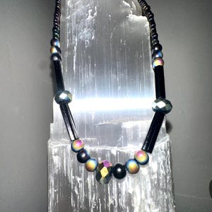 May include: A black beaded necklace with iridescent and faceted beads. The necklace features a mix of round and rectangular beads, with a central faceted bead. The beads have a metallic sheen, reflecting colors like purple, blue, and gold.