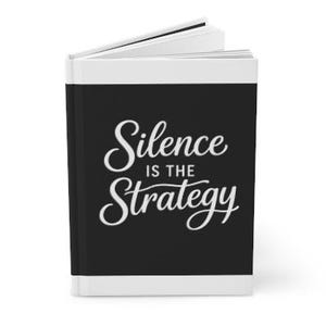 Peut inclure: Un journal à couverture rigide avec une couverture noire portant le texte blanc "Silence is the Strategy". Le journal a une bordure blanche et des pages blanches. Le livre est ouvert, révélant les pages intérieures.