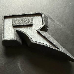 May include: A black, three-dimensional letter "R" emblem with a textured surface. The emblem has a rectangular top and a stylized, pointed bottom. The letter is set against a dark background.