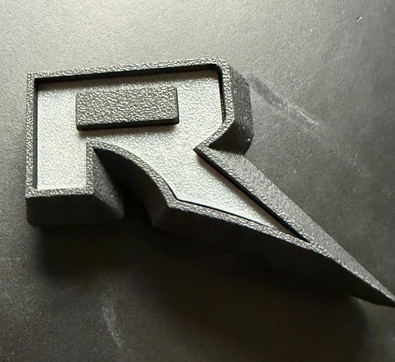 May include: A black, three-dimensional letter "R" emblem with a textured surface. The emblem has a rectangular top and a stylized, pointed bottom. The letter is set against a dark background.