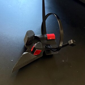May include: A black, 3D-printed object with a bird-like beak shape, featuring red accents. A black zip tie is looped around the object. The item is on a dark surface.