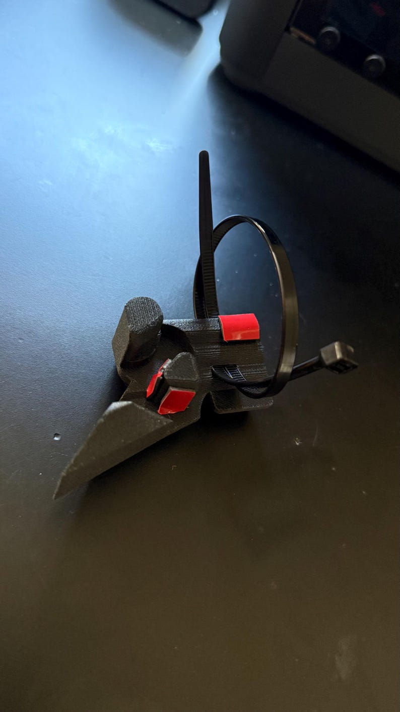 May include: A black, 3D-printed object with a bird-like beak shape, featuring red accents. A black zip tie is looped around the object. The item is on a dark surface.