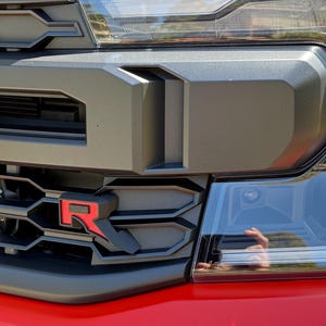 May include: Close-up of a black and red truck front end. The grill features a prominent "R" logo. Headlights and a bumper are visible. The truck's body is red.