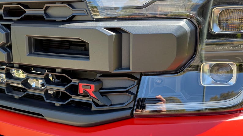 May include: Close-up of a black and red truck front end. The grill features a prominent "R" logo. Headlights and a bumper are visible. The truck's body is red.