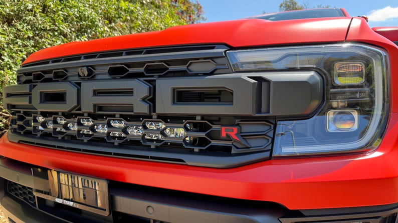 May include: Close-up of a red Ford Raptor truck front end. The black grille features the Ford name and a red "R" emblem. The headlights are large and angular, with a clear lens.
