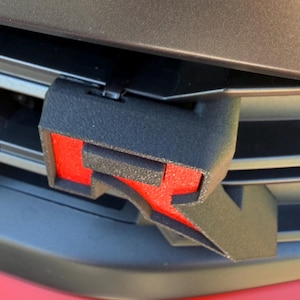 May include: Black and red car emblem featuring a stylized letter "R" on a black background. The emblem is mounted on a car's front grille, with a red bumper visible below.