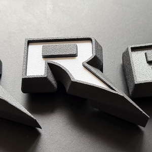 May include: Three gray letter "R" emblems with a textured finish. One has a blue interior, one has a white interior, and the third has a gray interior. The emblems are angled and appear to be 3D printed.