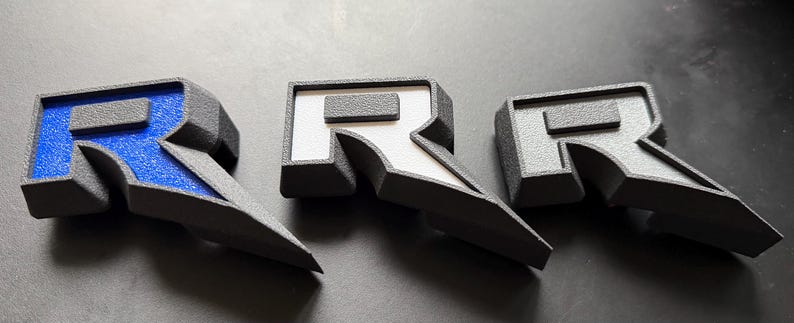 May include: Three gray letter "R" emblems with a textured finish. One has a blue interior, one has a white interior, and the third has a gray interior. The emblems are angled and appear to be 3D printed.