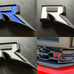 May include: A collection of automotive emblems featuring the letter "R" in various color schemes. The emblems have a textured finish and a stylized design with a sharp, angular element. One image shows the emblem installed on a vehicle's grille.