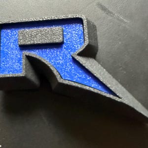 May include: A 3D-printed letter "R" in a stylized design. The letter features a textured black border with a vibrant blue interior. A small, rectangular black accent sits atop the "R". The design has a sharp, angular aesthetic.