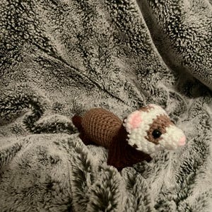 May include: A handmade crochet ferret toy in brown, white, and pink, resting on a soft, gray, faux fur blanket. The ferret has a detailed design with a white face, pink ears, and dark eyes.