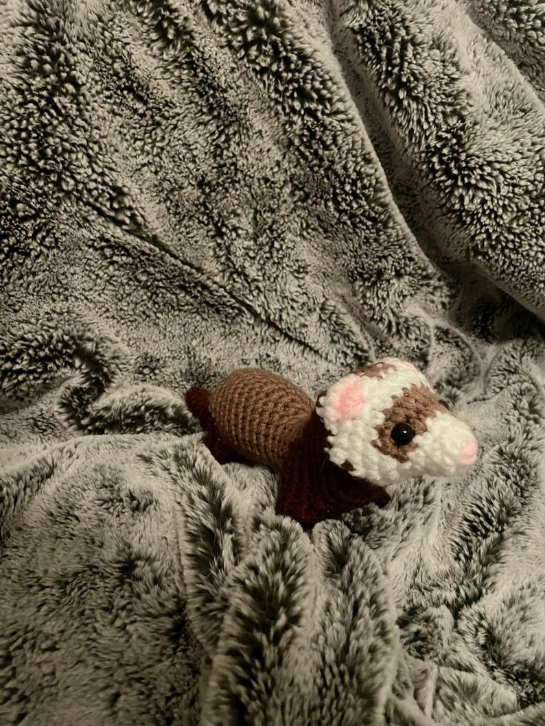 May include: A handmade crochet ferret toy in brown, white, and pink, resting on a soft, gray, faux fur blanket. The ferret has a detailed design with a white face, pink ears, and dark eyes.