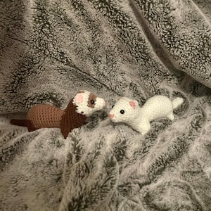 May include: Two crocheted ferret toys. One is brown with a white face and the other is white with pink accents. They are lying on a soft, gray, textured blanket. The toys are handmade and appear to be stuffed.
