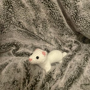 May include: A white crocheted ferret with black eyes and pink accents on its ears and nose rests on a gray, plush blanket. The ferret is handmade and has a detailed, textured appearance, showcasing the craftsmanship of the crochet work.
