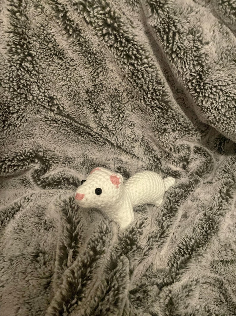 May include: A white crocheted ferret with black eyes and pink accents on its ears and nose rests on a gray, plush blanket. The ferret is handmade and has a detailed, textured appearance, showcasing the craftsmanship of the crochet work.