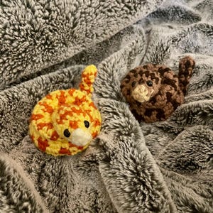 Pode incluir: Dois brinquedos de crochê em forma de cobra. Um é amarelo e laranja, o outro é marrom com manchas marrons mais escuras. Ambos têm pequenos detalhes brancos e pretos para os olhos e a boca. As cobras estão enroladas em uma superfície cinza texturizada.