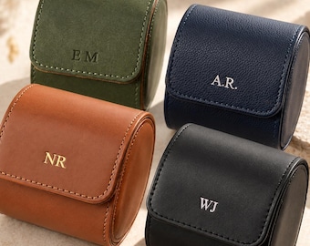 May include: Four cylindrical watch cases in olive green, navy blue, tan, and black. Each case features a flap closure with a personalised monogram: EM, A.R., NR, and WJ. The cases have a smooth or textured finish and visible stitching along the edges.