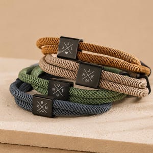 May include: A stack of four braided cord bracelets in mustard yellow, beige, sage green, and slate blue. Each bracelet features a rectangular black metal clasp engraved with a unique cross design containing letters, numbers, and small symbols.