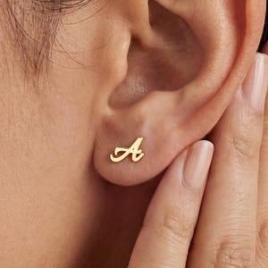 May include: A close-up of a gold-colored stud earring in the shape of the letter "A" on a person's ear. The earring is small and delicate, with a cursive font. The person's skin tone is light.