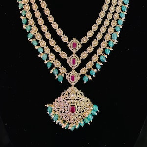 May include: A gold-toned jewelry set featuring a multi-strand necklace and matching earrings. The necklace has a pendant with a red gemstone and is adorned with green beads and white pearls. The earrings also have red gemstones and green beads.