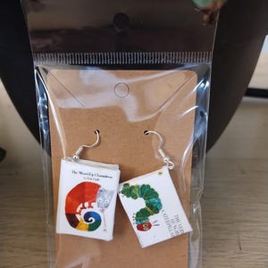 May include: A pair of silver-tone dangle earrings featuring miniature book covers. One earring displays "The Mixed-Up Chameleon" by Eric Carle, the other "The Very Hungry Caterpillar." The earrings are packaged on a brown card within a clear plastic bag.