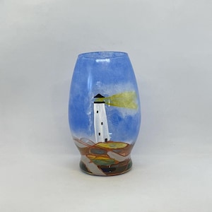 May include: A hand-painted glass vase with a blue and white lighthouse scene. The lighthouse is white with a black roof and a yellow beam of light. The vase has a brown, yellow, and green base.