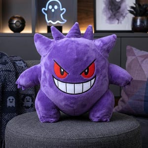 Könnte beinhalten: Ein lila Gengar-Plüschtier mit roten Augen und einem breiten Grinsen. Das Stofftier hat stachelige Auswüchse auf Kopf und Armen. Es sitzt auf einem grauen Hocker, im Hintergrund ist ein geisterförmiges Neonlicht zu sehen.