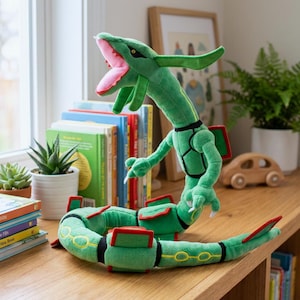 Könnte beinhalten: Ein grünes Plüschdrachen-Spielzeug mit einem langen Körper und einem geöffneten Maul, das rosa und rote Details zeigt. Der Drache hat gelbe Akzente und rote Flossen. Es liegt auf einer Holzoberfläche in der Nähe von Büchern und einer kleinen Pflanze.