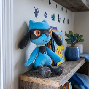 May include: A plush toy of the character Lucario, featuring light blue and dark grey fur, orange eyes, and a black mask-like pattern. The stuffed animal has long ears, a small white collar, and sits upright with its limbs extended forward.
