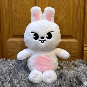 以下が含まれることがあります： ピンク色の内側の耳とピンク色のお腹を持つ、白いウサギのぬいぐるみ。ウサギは大きな黒い目、小さな黒い口、そして遊び心のある表情をしています。灰色のラグの上に座っています。
