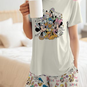 May include: A light-colored pajama set featuring a graphic of Disney characters, including Mickey and Minnie Mouse, Donald and Daisy Duck, and Pluto. The shorts and t-shirt are decorated with Easter-themed designs and the words "Happy Easter".