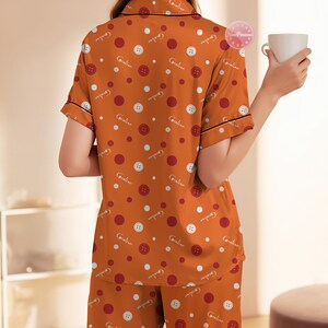 May include: Orange pajama set with a button and polka dot pattern. The short-sleeved top and pants feature a repeating design of white buttons, red and white polka dots, and the word "Caroline". Black trim accents the collar and sleeves.