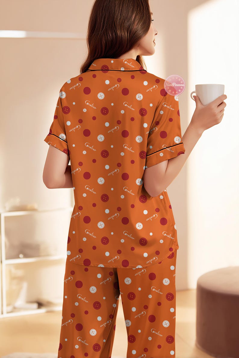 May include: Orange pajama set with a button and polka dot pattern. The short-sleeved top and pants feature a repeating design of white buttons, red and white polka dots, and the word "Caroline". Black trim accents the collar and sleeves.
